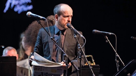Der Musikkünstler Matthew Herbert steht mit einer Zeitung auf der Bühne und spricht in ein Mikrofon. © picture alliance / abaca 